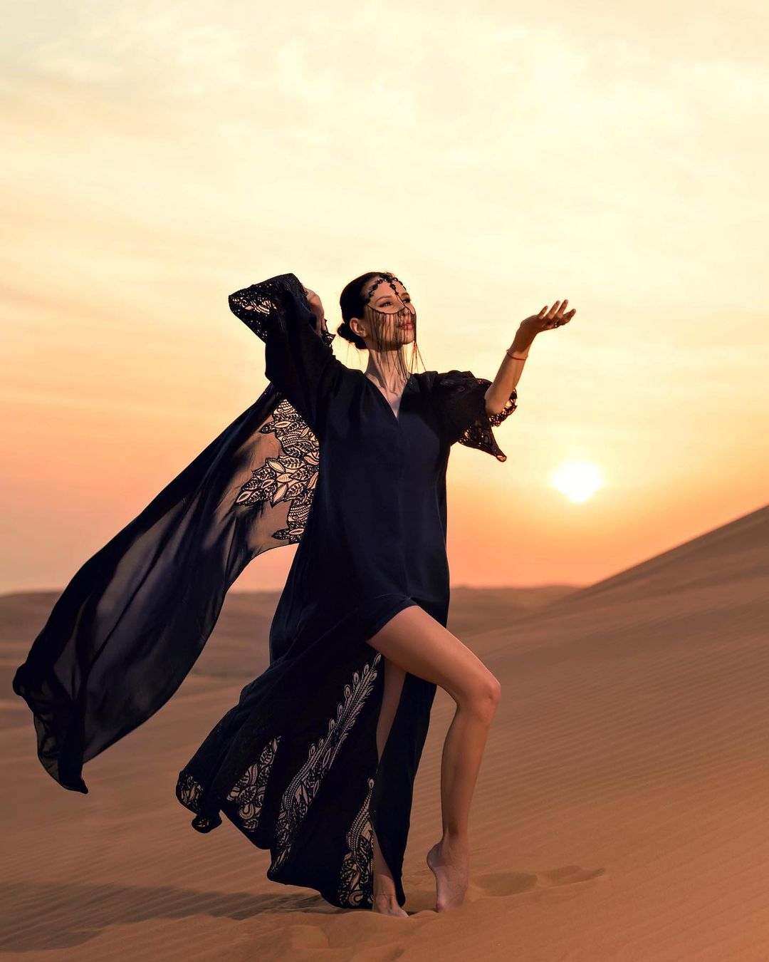 A woman in black abaya and niqab poses alone in the Dubai desert, standing on a sand dune overlooking the vast empty desert landscape.