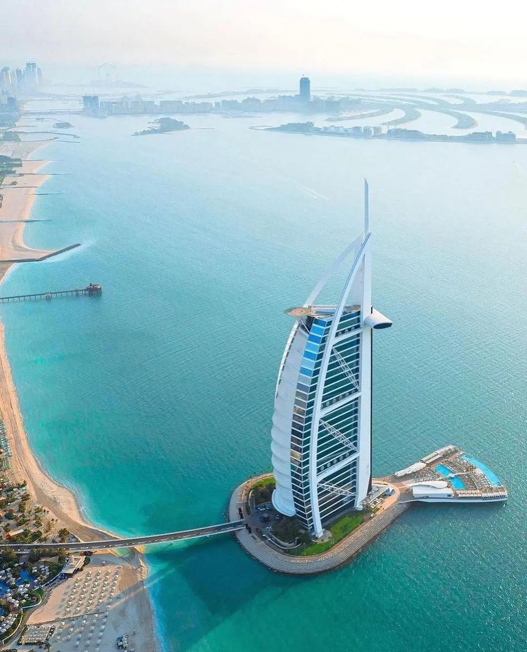 An aerial view of the iconic Burj Al Arab luxury hotel on its own man-made island in Dubai, its distinctive sail-shaped silhouette towering over the coastline at sunrise.