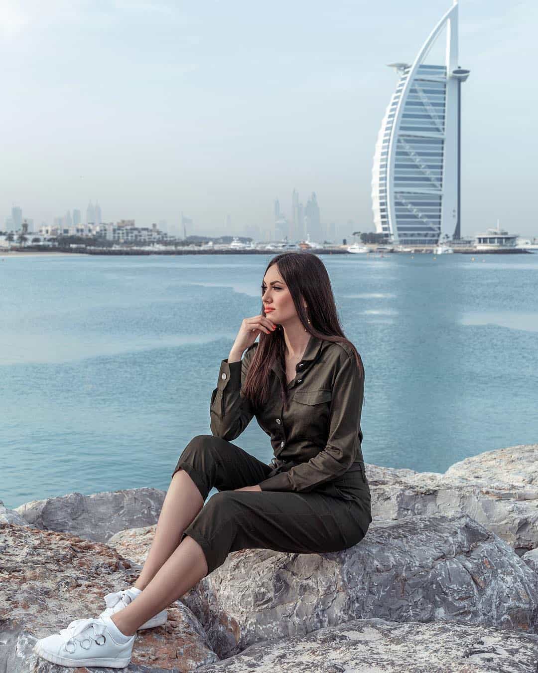 Красивая девушка сидит на камнях на побережье Дубая около пятизвездочного отеля Бурж аль Араб.