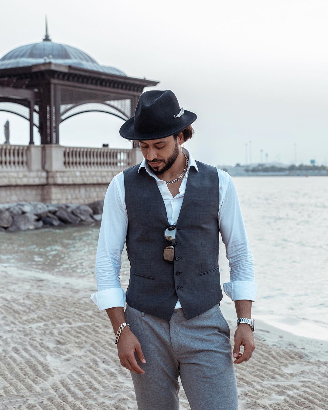 An Arab man wearing smart casual European clothing poses with a serious expression on a beach in Abu Dhabi with the city skyline behind him.