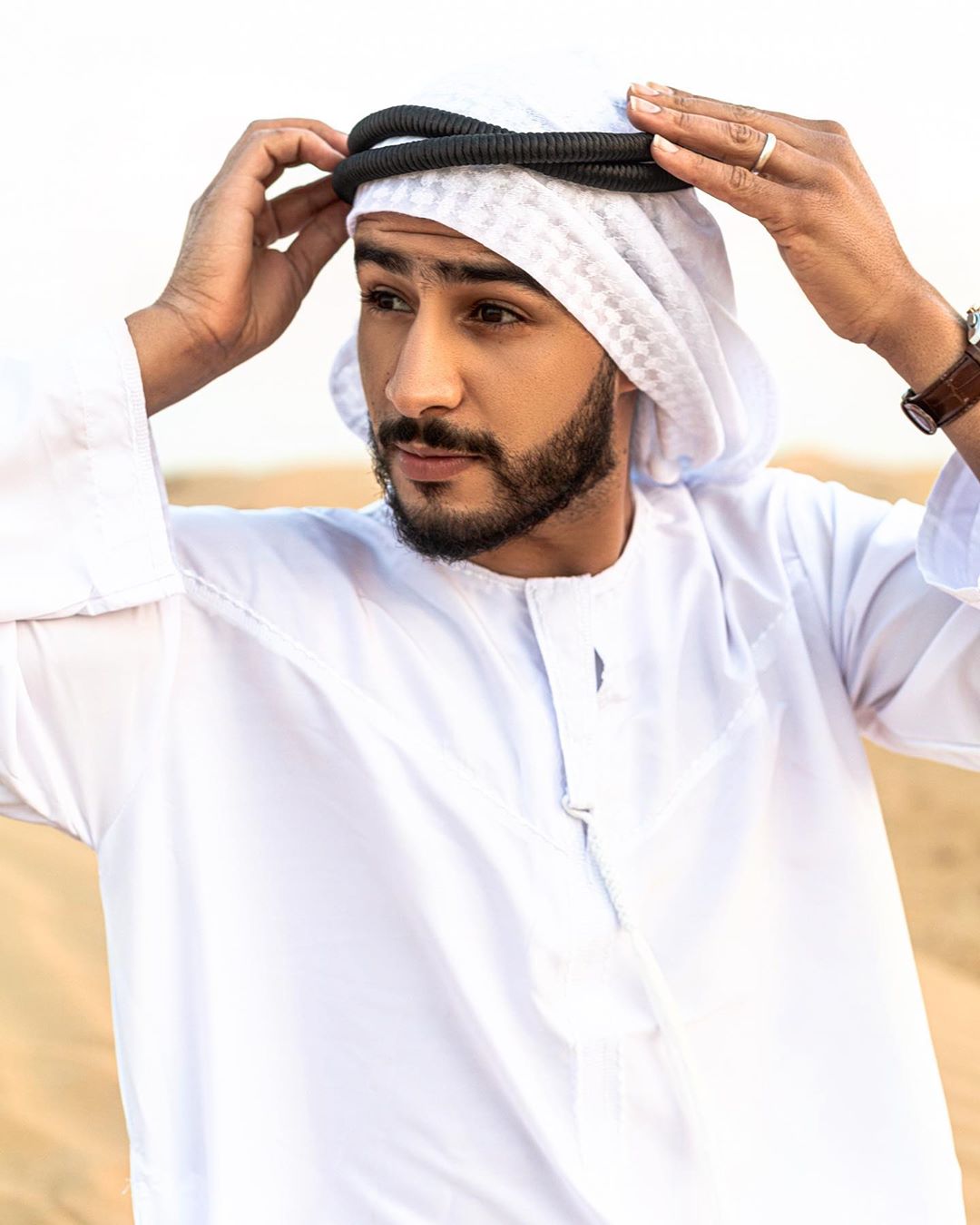 A young Arab man with a trimmed beard poses in traditional Emirati attire, wearing a white kandura robe and ghutra headdress, against a neutral desert background.
