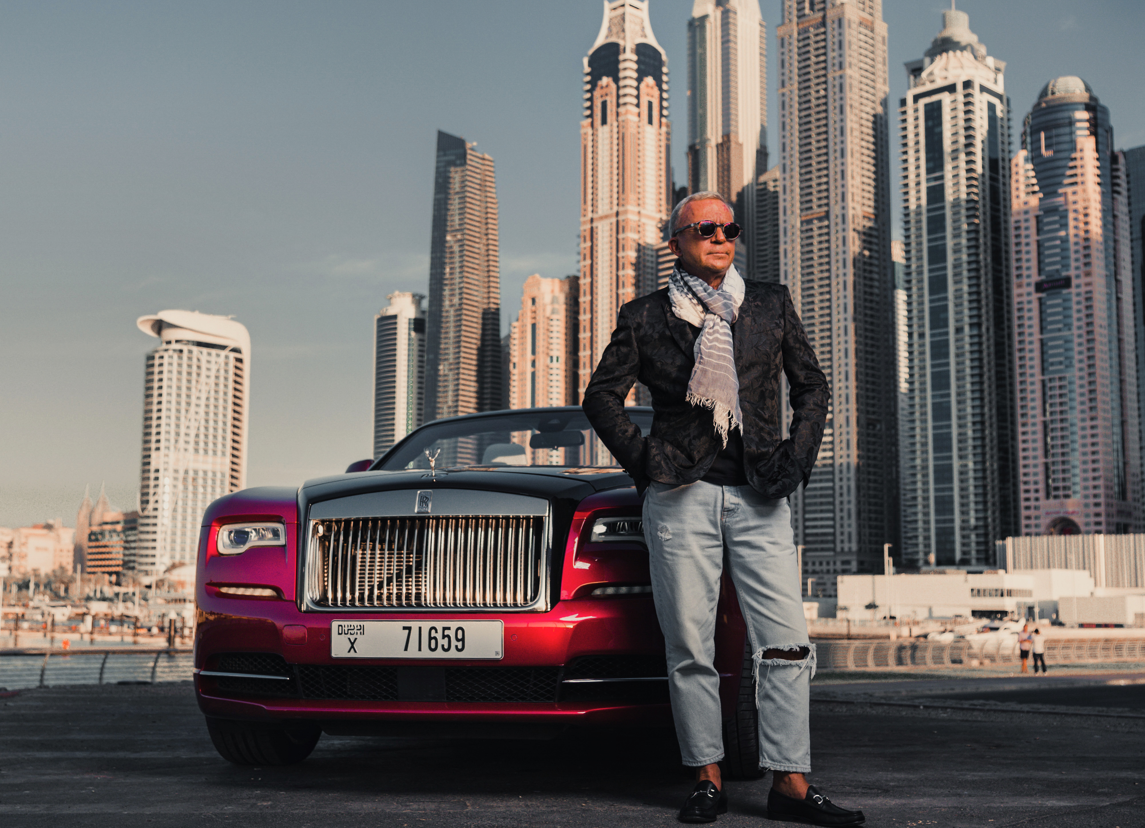 A man is posing next to a luxurious red Rolls-Royce during a photo shoot in Dubai Marina.